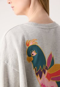 Grijze sweater met een levendig geborduurd vogelontwerp in teal, roze en geel op de achterkant. Zachte textuur, casual pasvorm.