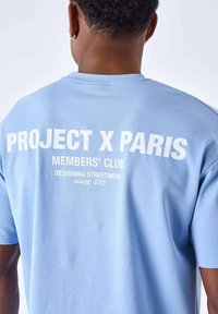 Lichtblauw katoenen t-shirt met op de achterkant witte bedrukte tekst: "PROJECT X PARIS MEMBERS' CLUB DESIGNING STREETWEAR sinds 2015."