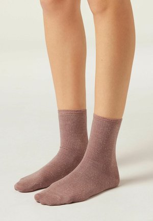 Une paire de jambes portant des chaussettes mauves mi-mollet avec un motif léger de points, se tenant sur une surface unie de couleur claire.