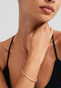 Bracciale bianco sottile con piccoli accenti dorati, indossato su un polso abbronzato; sullo sfondo si vede un muro grigio texture.