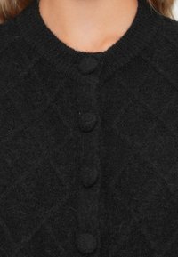 Svart stickad cardigan med ett strukturerat diamantmönster, rund hals och fem runda knappar. Mjuk, mysig tyg med lätt fluffighet.