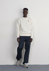 Sudadera crema con un pequeño logo, combinada con pantalones oscuros y zapatillas grises. La tela tiene una textura suave y presenta un corte relajado.
