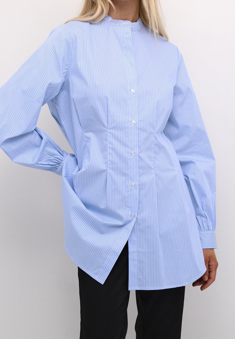 Ljusblå, randig skjorta med knappar och hög krage samt långa ärmar. Har en rundad nederkant och lätt samling vid axlarna.