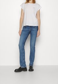 Camiseta blanca de manga corta, jeans de mezclilla azul, corte ajustado, con detalles de costura visibles, combinados con zapatos negros gruesos.