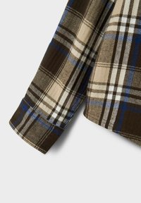 Polsino a manica lunga di una camicia in flanella a quadri marrone, beige e blu, con cuciture dettagliate su sfondo grigio chiaro.