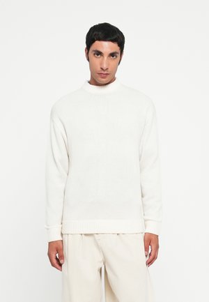 Suéter blanco de punto con un cuello alto, puños acanalados y dobladillo. Combinado con pantalones beige claros de corte relajado. Textura suave.