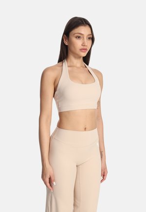 Ung kvinde iført matchende beige halterneck crop top og høj talje leggings, stående mod en ensfarvet lys baggrund.