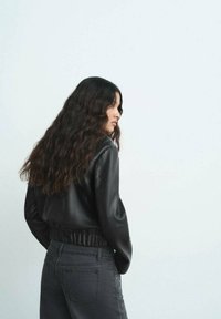 Giacca in finta pelle nera con vita elastica, abbinata a jeans grigi scuri con dettagli laterali. Capelli ondulati ricadono sulle spalle.