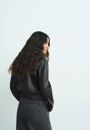 Giacca in finta pelle nera con vita elastica, abbinata a jeans grigi scuri con dettagli laterali. Capelli ondulati ricadono sulle spalle.