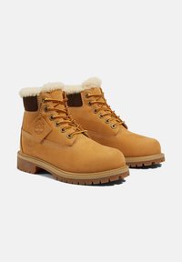 Tanfarbene Stiefel mit einem Fellfutter am Schaft, ausgestattet mit braunen Akzenten, metallischen Ösen, gelben Schnürsenkeln und einer robusten Gummisohle.