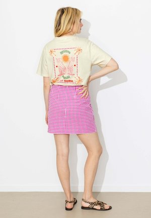 Femme portant un T-shirt crème avec un motif soleil et léopard et une jupe vichy rose, debout, une main sur la hanche, tournée dos à la caméra.