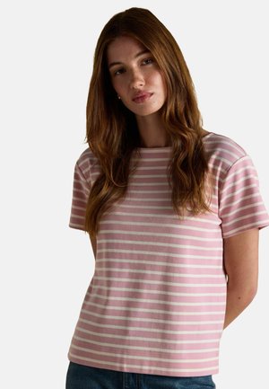 Junge Frau mit langen braunen Haaren, die ein kurzärmliges, gestreiftes Shirt in Rosa und Weiß sowie eine blaue Jeans trägt, vor einem schlichten Hintergrund.