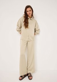 Camicia button-up in satin beige con spalle scese, abbinata a pantaloni a gamba larga. Indossata con sandali marrone scuro con doppie cinghie.
