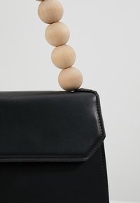 Sac à main noir avec rabat géométrique et poignée composée de grosses perles rondes en bois clair, posé sur un fond uni clair.