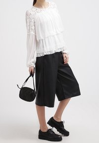 Blouse blanche à couches avec accents en dentelle, associée à un pantalon culotte plissé noir. Les accessoires comprennent un petit sac à bandoulière noir et des chaussures noires à lacets.