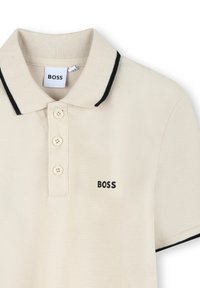 Krémová polo košela s čiernym lemovaním goliera, trojknoflíkovou légou a vyšitým logom "BOSS" na ľavej hrudi v čiernej farbe. Textúrovaná látka.