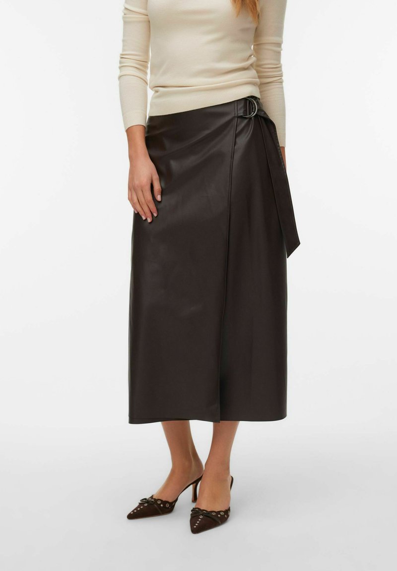 Kvinde iført en beige langærmet top, mørkebrun læder midi slå-om nederdel med spænde og mørkebrun spids tå hæle med udskæringsdetaljer.