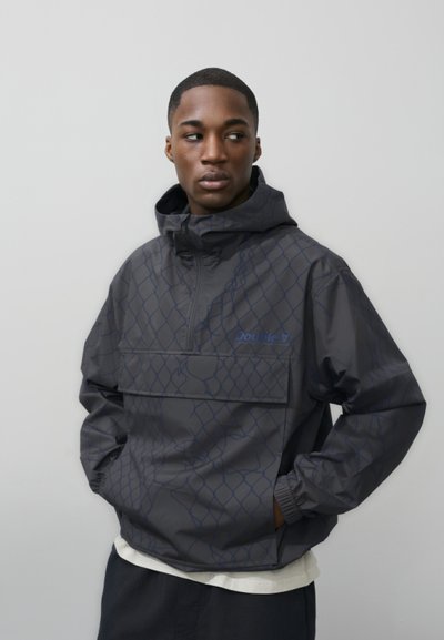 Jeune homme portant une veste à capuche gris foncé à motif, les mains dans les poches, regardant sur le côté devant un fond gris uni.