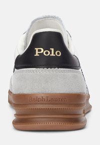 Sneakers i vitt läder och gråmocka, svart läderhalk med guld "Polo"-logotyp, och brun gummisula med texturerad finish.