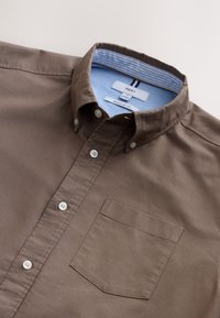 Camisa marrón abotonada con un corte regular, que presenta un cuello puntiagudo, botones blancos y un bolsillo en el pecho. El interior del cuello es de rayas azul claro.