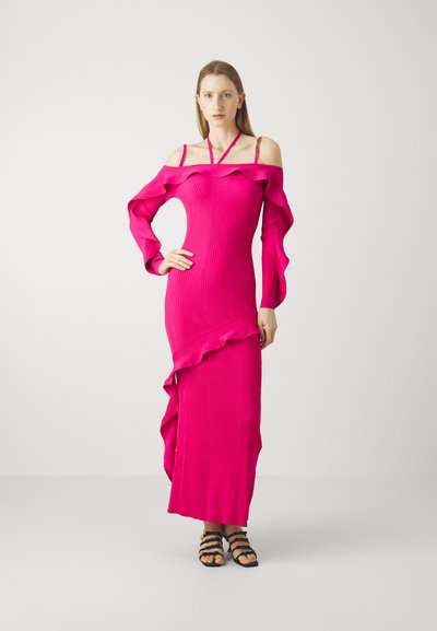 Robe longue et ajustée en rose vif avec des manches décolletées et des accents de volants. Associée à des sandales noires à lanières.