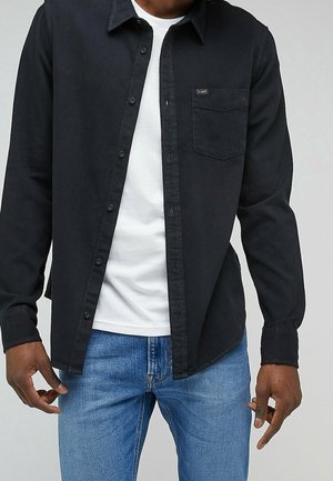 Mann mit offenem schwarzem Langarmhemd über weißem T-Shirt und blauer Jeans, stehend vor einem schlichten hellgrauen Hintergrund.