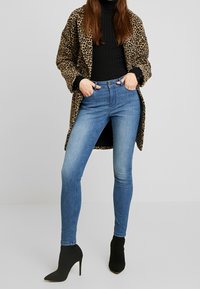 Manteau en imprimé léopard sur un col roulé noir, associé à un jean skinny bleu et des bottines noires à talons pointus. Texture subtile des tissus visible.