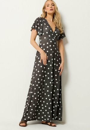Kobieta z długimi blond włosami ubrana w czarną sukienkę maxi na krótki rękaw w białe kropki, stojąca na tle gładkiej białej ściany.