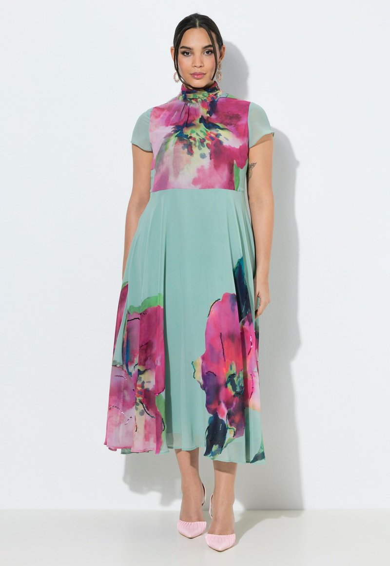 Vrouw draagt een midi-jurk met hoge hals en korte mouwen in lichtgroen met grote roze en paarse bloemenprints, gecombineerd met roze puntige hakken.