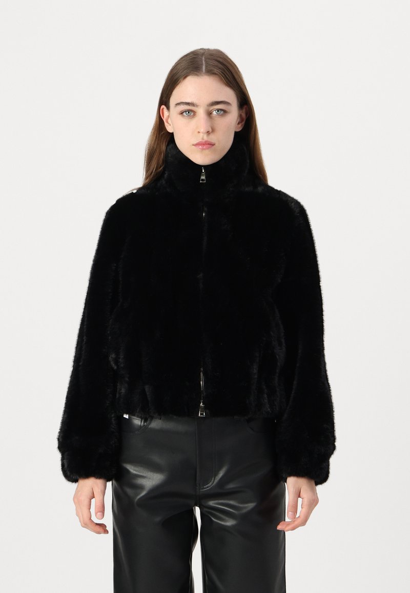 Schwarze Kunstpelzjacke mit hohem Kragen, Reißverschluss und weit geschnittenen Ärmeln. Strukturiertes Finish, verkürztes Design, getragen über schwarzen Lederhosen.