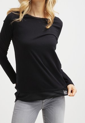 Femme portant un haut noir à manches longues avec un ourlet transparent, associé à un jean gris clair, montrée du cou aux hanches sur un fond blanc.