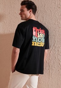 Černé tričko s krátkým rukávem, na zadní straně se nachází vícebarevný nápis "tohle není nové" na texturovaném pozadí.