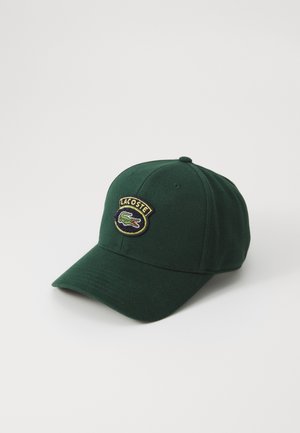 Grønn baseballcaps laget av bomull, med et brodert Lacoste-logo i en sirkulær lapp på fronten. Buet skjerm, teksturert stoff.