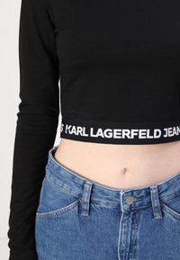 Crni dugih rukava crop top s bijelom logotip trakom, uparen s trapericama visokog struka u plavoj boji koje imaju gumb i džepove.