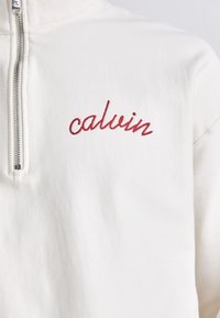 Sudadera blanca con cremallera que presenta el logotipo "calvin" bordado en rojo en el lado izquierdo del pecho. Fabricada con una suave tela texturizada y con un cuello alto.