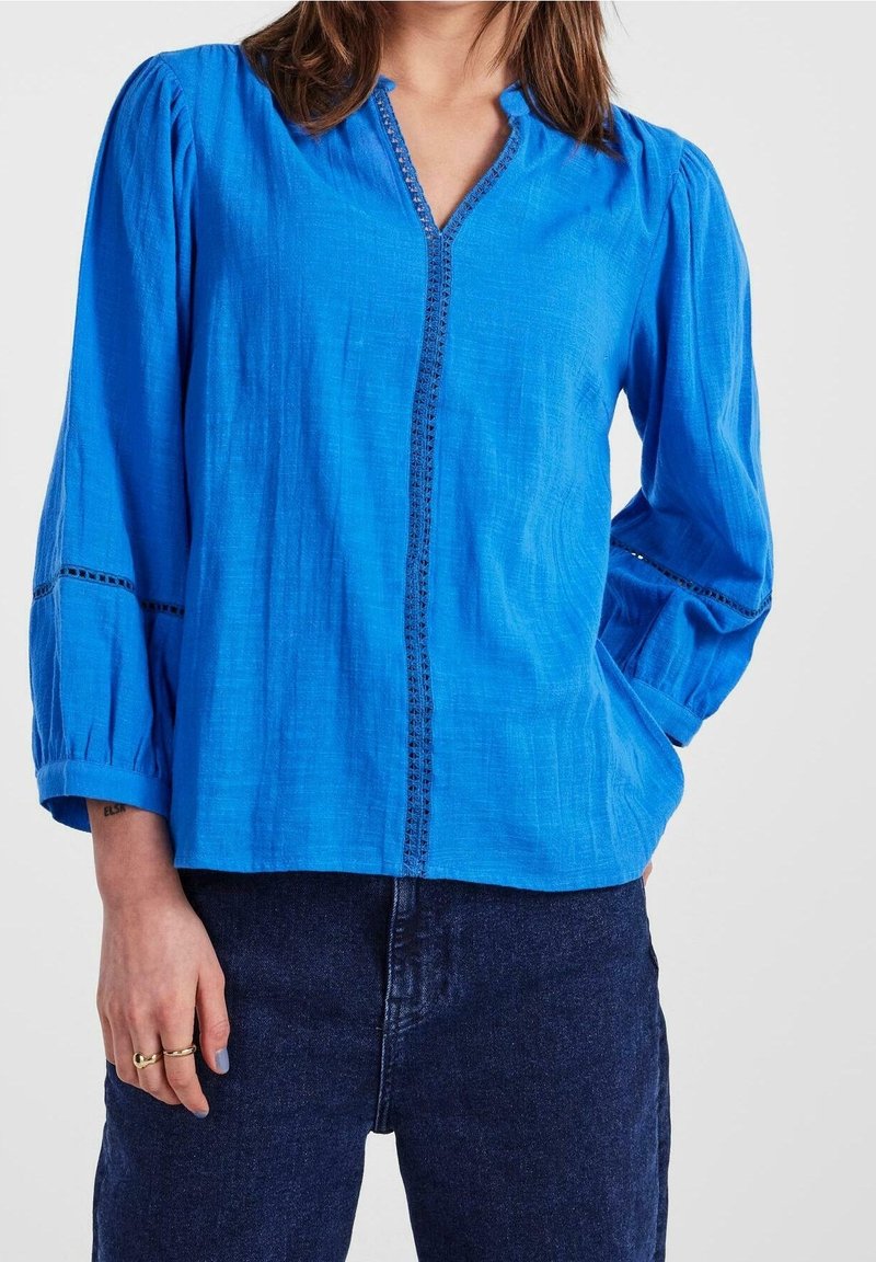 Blouse légère bleue avec un col en V et patte de boutonnage, dotée de manches froncées et de coutures décoratives le long des coutures. Portée avec un jean foncé.