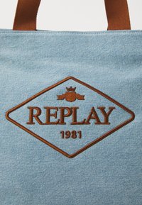 Denim tote taske i lyseblå med brune læderdetaljer. Har et diamantformet logo med "REPLAY" og "1981" broderet i brunt.