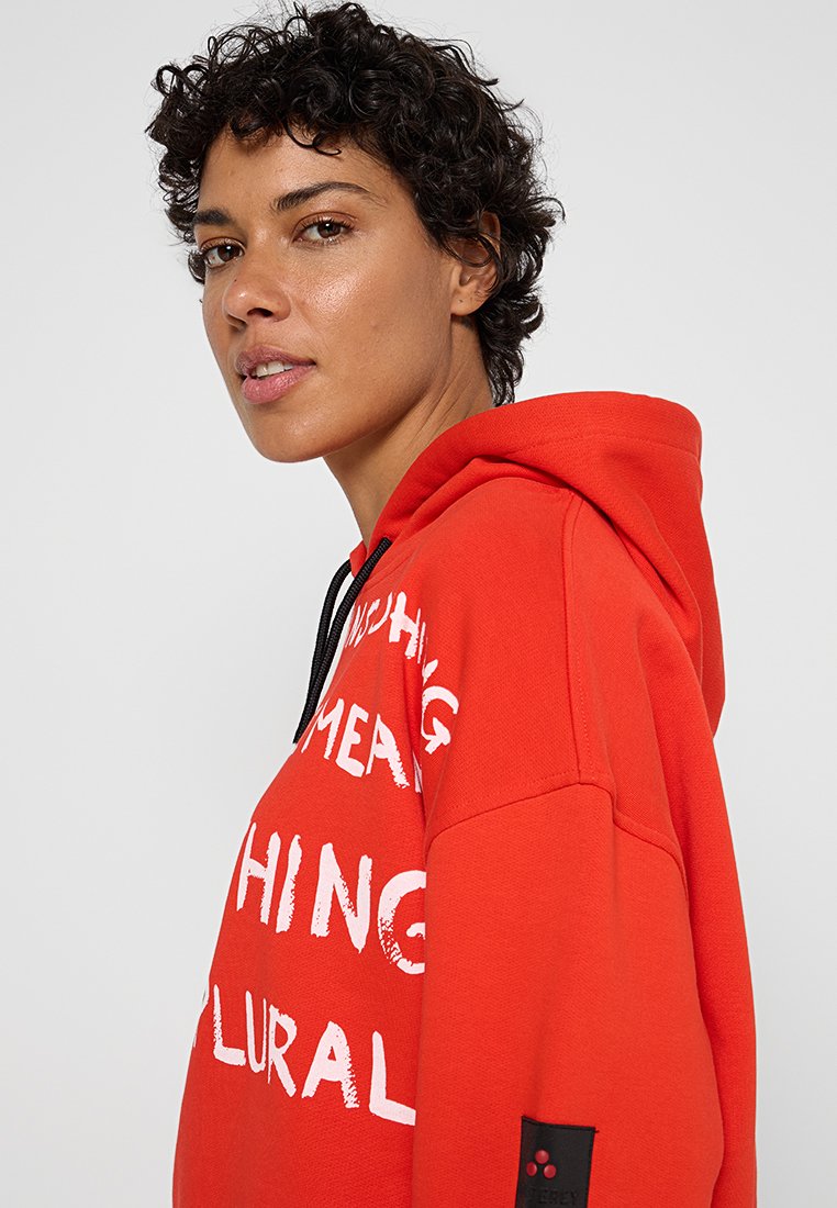 Kobieta z krótkimi, kręconymi włosami, ubrana w jasnoczerwoną bluzę z kapturem, z białym nadrukiem i czarnymi sznurkami, zwrócona bokiem w kierunku szarego tła.