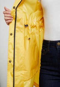 Geel regenjas met een binnenvoering, trekkoord, rits- en drukknoopsluitingen, en een zijzak. De buitenstof is glad en lichtgewicht.