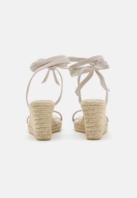 Beige espadrille-kiler med en flettebunden jute hæl, med bløde stofremmer som bindes ved ankelen. Glat tekstur og åben rygdesign.