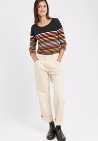 Gestreiftes Langarmshirt in Schwarz, Rot, Gelb und Weiß, kombiniert mit cremefarbenen Hosen mit umgeschlagenen Bündchen und schwarzen Knöchelschuhen.