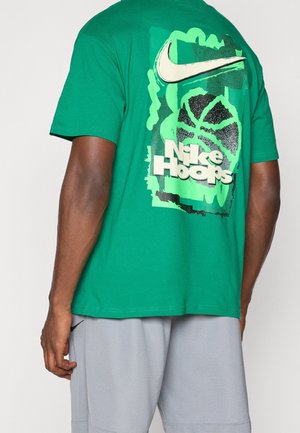 Mann trägt ein grünes Nike Hoops T-Shirt mit Basketballgrafik und weißem Nike-Swoosh auf dem Rücken, kombiniert mit hellgrauen Shorts.