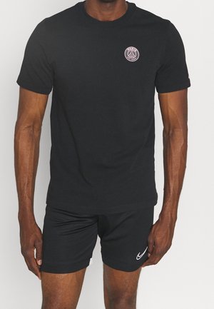 Mann trägt ein schwarzes Paris Saint-Germain T-Shirt und schwarze Nike-Shorts und steht vor einem einfarbigen Hintergrund.