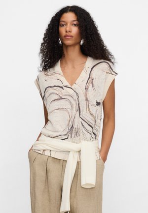 Kobieta z długimi kręconymi włosami, ubrana w bezrękawnik z abstrakcyjnymi beżowo-brązowymi wzorami, beżowe spodnie oraz związaną lekką sweterek wokół talii.