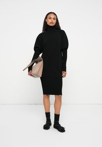 Μαύρο ριγωτό πλεκτό φόρεμα με ψηλό γιακά, στενό φούστα και φαρδιές μανσέτες. Το μοντέλο κρατά μια μπεζ τσάντα. Φορά μαύρες κάλτσες και σανδάλια.