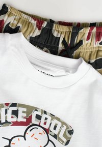 Witte T-shirt met een ronde halslijn en een grafische print. Hieronder camouflagetrouser met een groene, rode en zwarte print en een elastische tailleband.