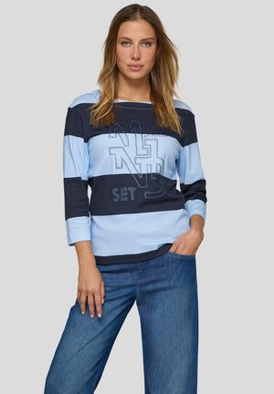 Frau trägt ein langärmeliges Shirt mit hellblauen und dunkelblauen Streifen und dem Text "N1ND SET", kombiniert mit blauen Jeans, stehend vor einem grauen Hintergrund.