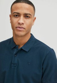 Polo-shirt bleu marine en tissu texturé, avec deux boutons au col et un petit logo sur la poitrine gauche. Design lisse et bien ajusté.