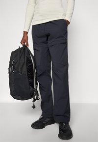 Pantalons noirs d'extérieur avec poches zippées, associés à un sac à dos noir avec un tissu texturé et des bretelles ajustables. Chaussures de sport noires.