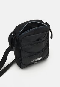 Borsa a tracolla nera con molte tasche con zip, tessuto testurizzato, tracolla regolabile e logo del marchio visibile sul davanti.